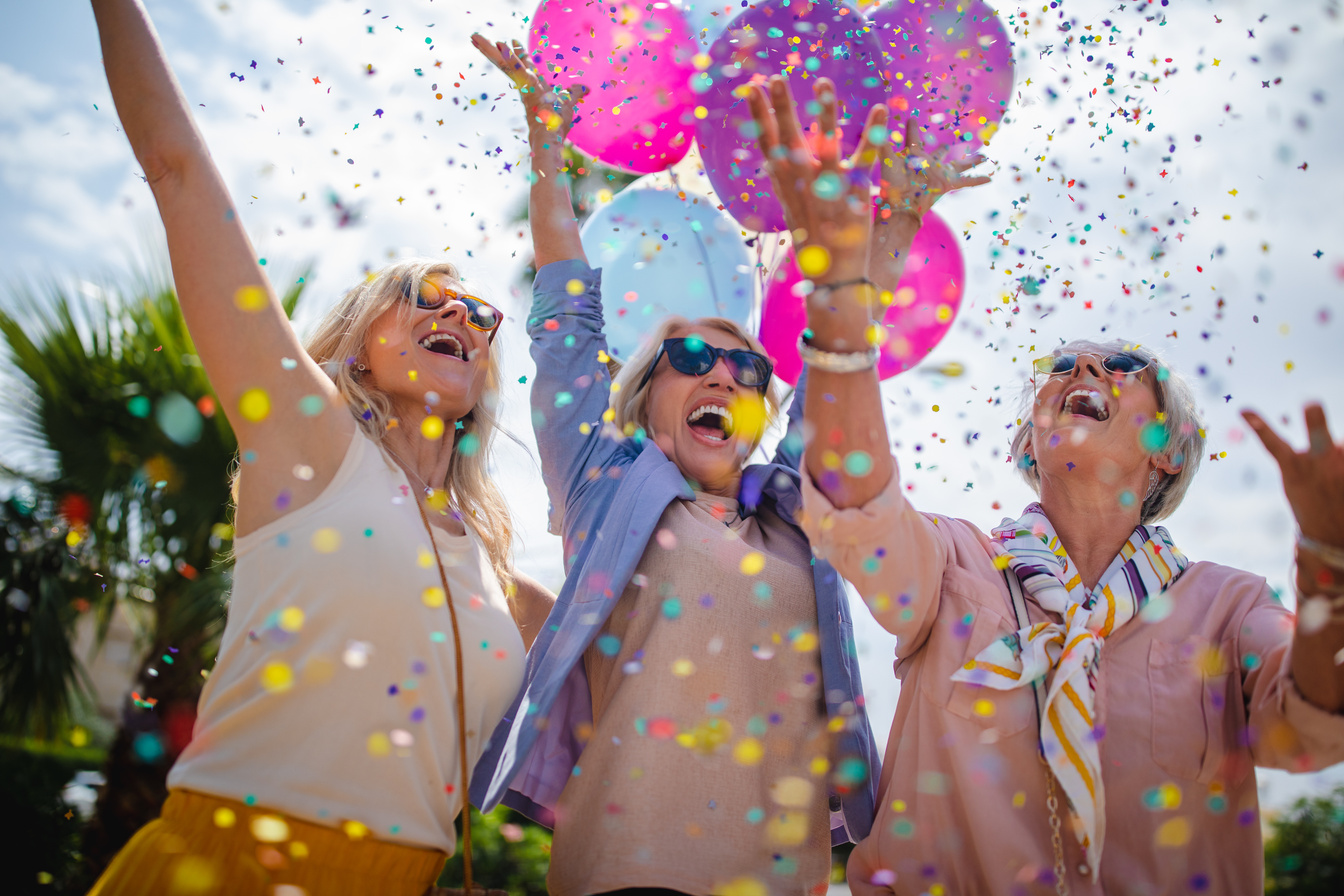 Excited mature women celebrating with colorful confetti and balloons outdoors